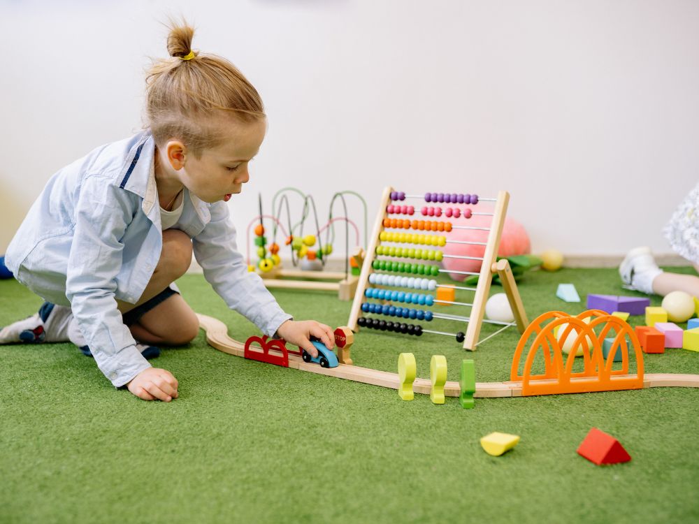 Criança brincando no chão com trilho de madeira, carrinho e brinquedos educativos coloridos, incluindo ábaco e peças de encaixe em ambiente infantil.